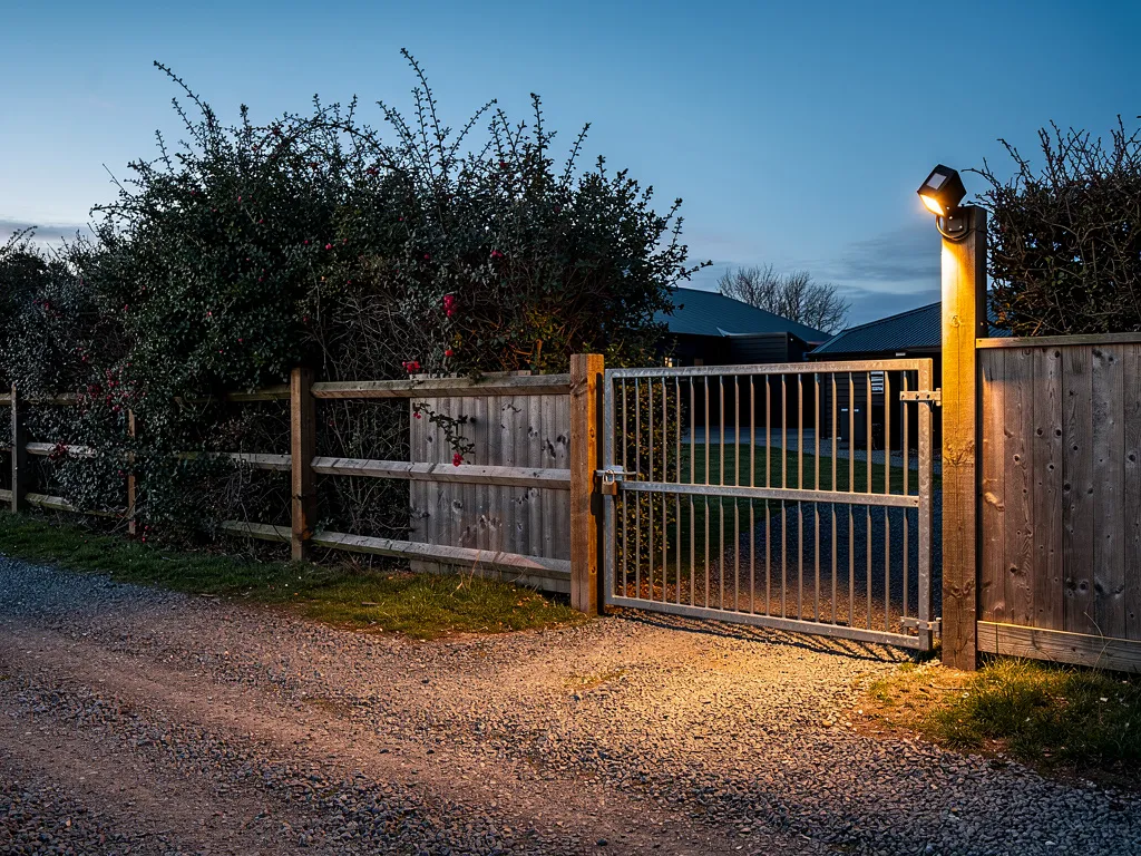 Rural homestead perimeter with wooden fence, motion-sensor lighting, and reinforced gate at dusk