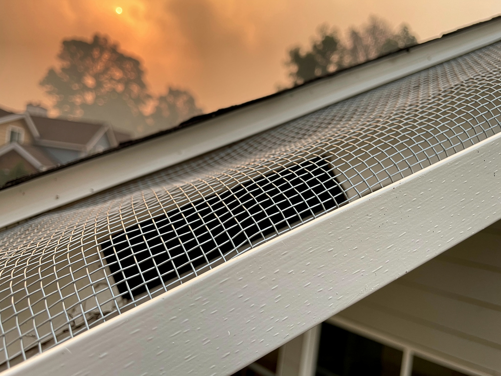 Ember-resistant 1/8-inch mesh vent screens installed on a house soffit with wildfire smoke visible in the background, showing the barrier that blocks ember intrusion into the attic